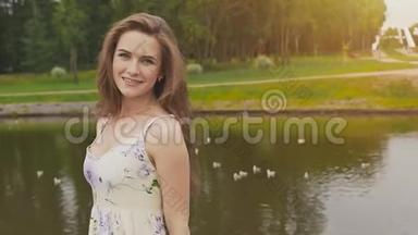 美丽的年轻女孩，留着长发，穿着一件夏装，在公园一个池塘的背景上对着镜头摆姿势。 帆布